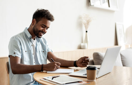 A person writes in a notebook sitting at a desk in front of a laptop computer
